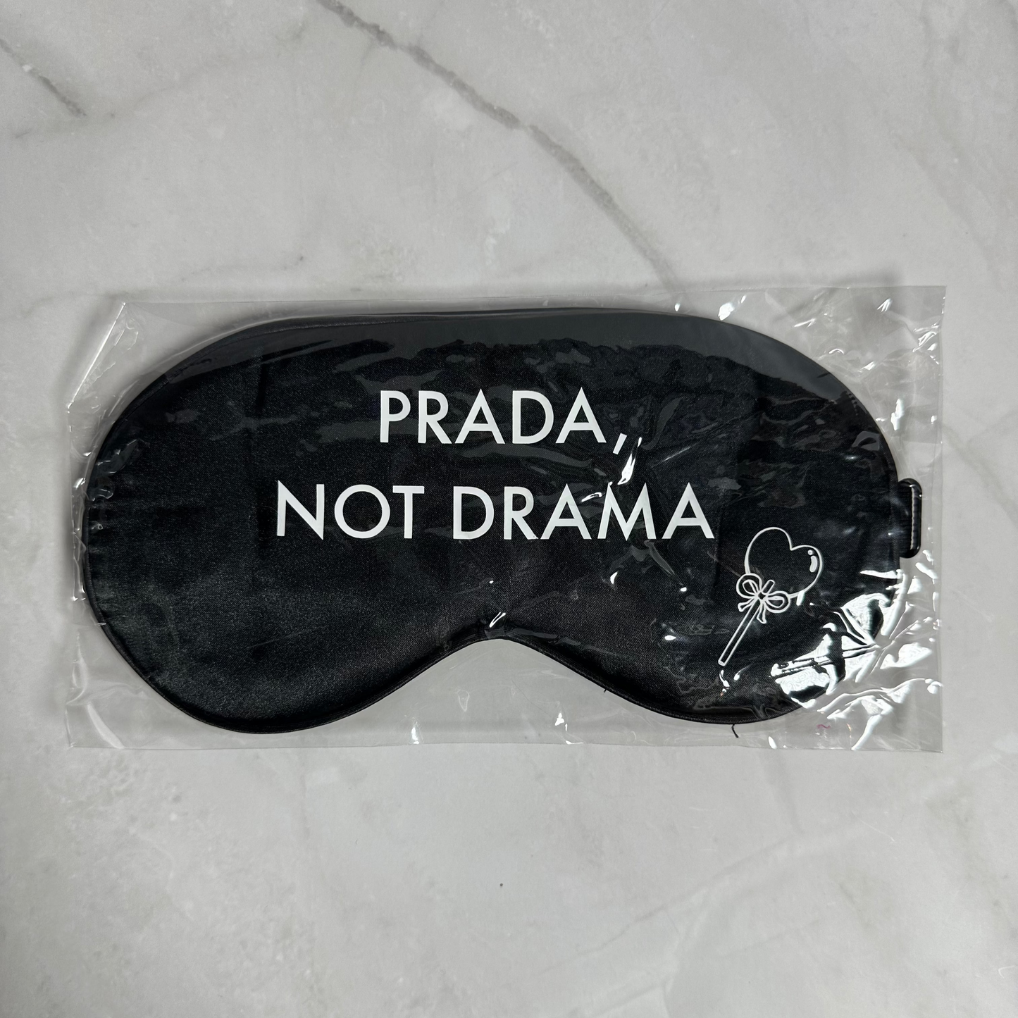 Black sleep mask with 'Prada Not Drama' text in clear packaging on a marble surface