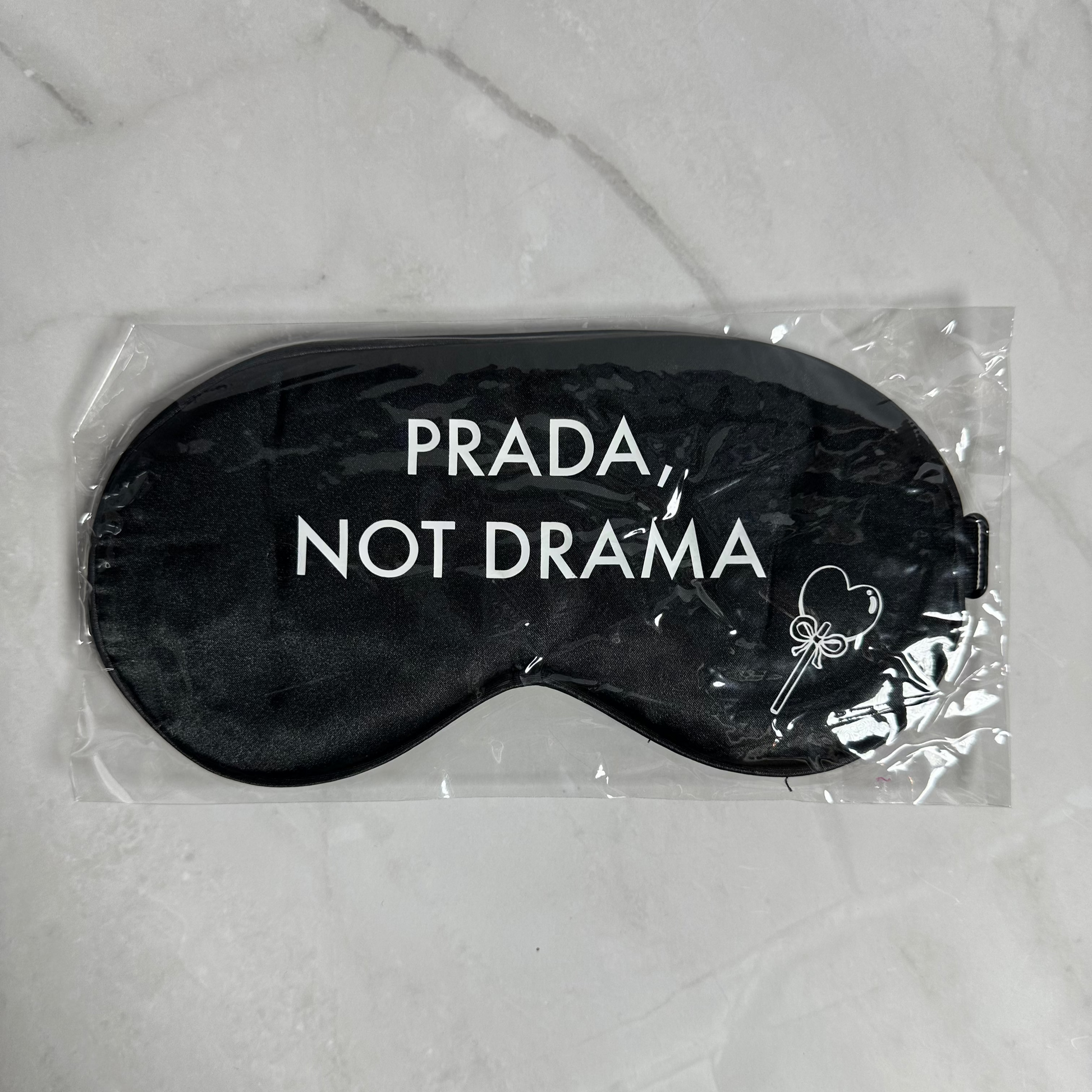 Black sleep mask with 'Prada Not Drama' text in clear packaging on a marble surface
